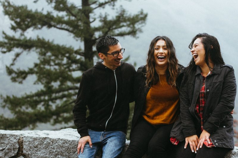 Photo of one man and two women sat on a stone wall laughing and talking about happiness self esteem